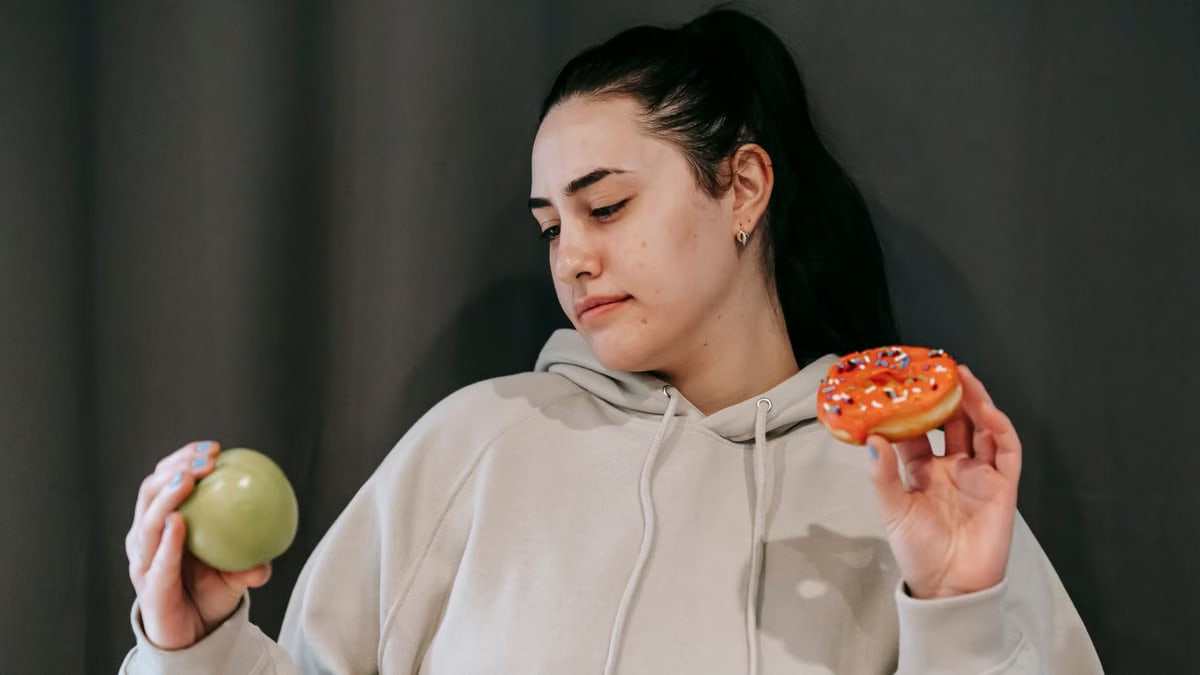 Pensive female wearing casual hoodie deciding between healthy green apple and sweet doughnut in studio